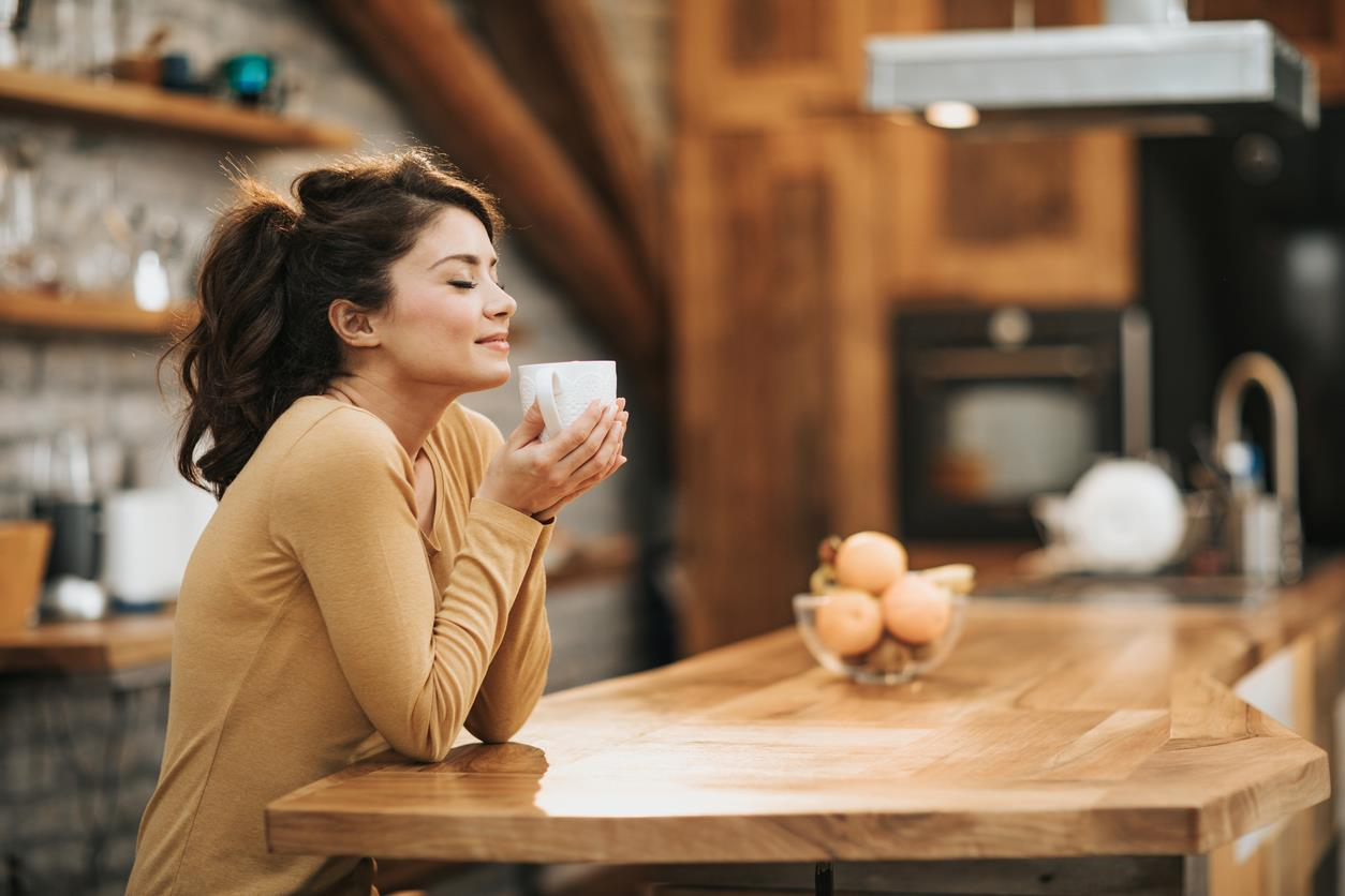 Woman at a kitchen counter with a mug in hand, closing her eyes and grinning. 